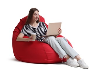 Beautiful young woman with laptop and paper cup of drink having online meeting while sitting on red bean bag chair against white background