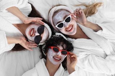 Photo of Happy friends with facial masks and sunglasses on bed, top view. Spa party