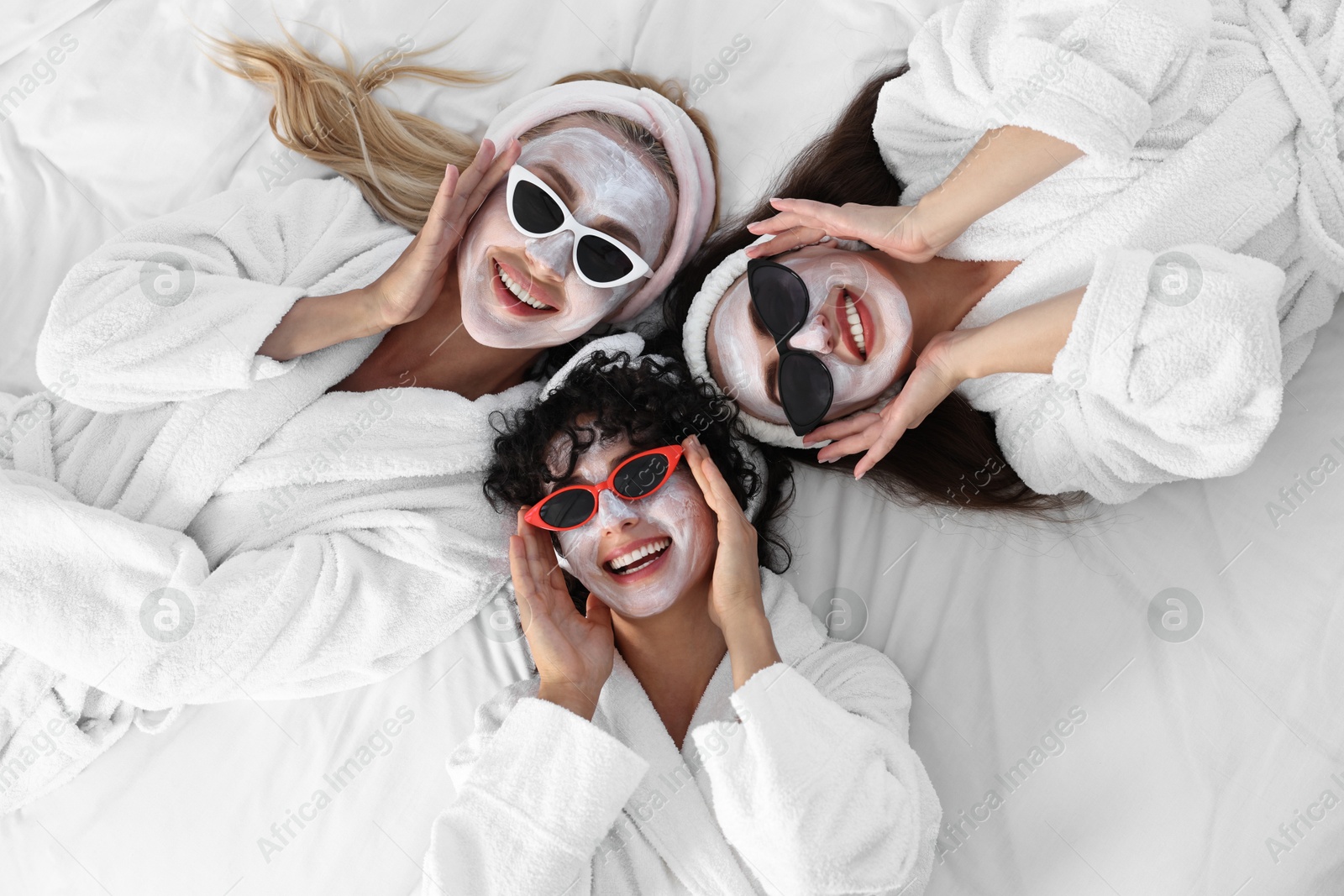 Photo of Happy friends with facial masks and sunglasses on bed, top view. Spa party