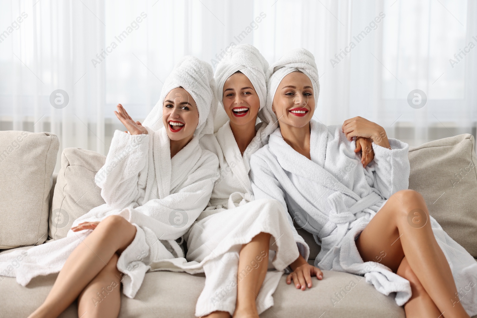 Photo of Happy friends in bathrobes with towels on couch indoors. Spa party