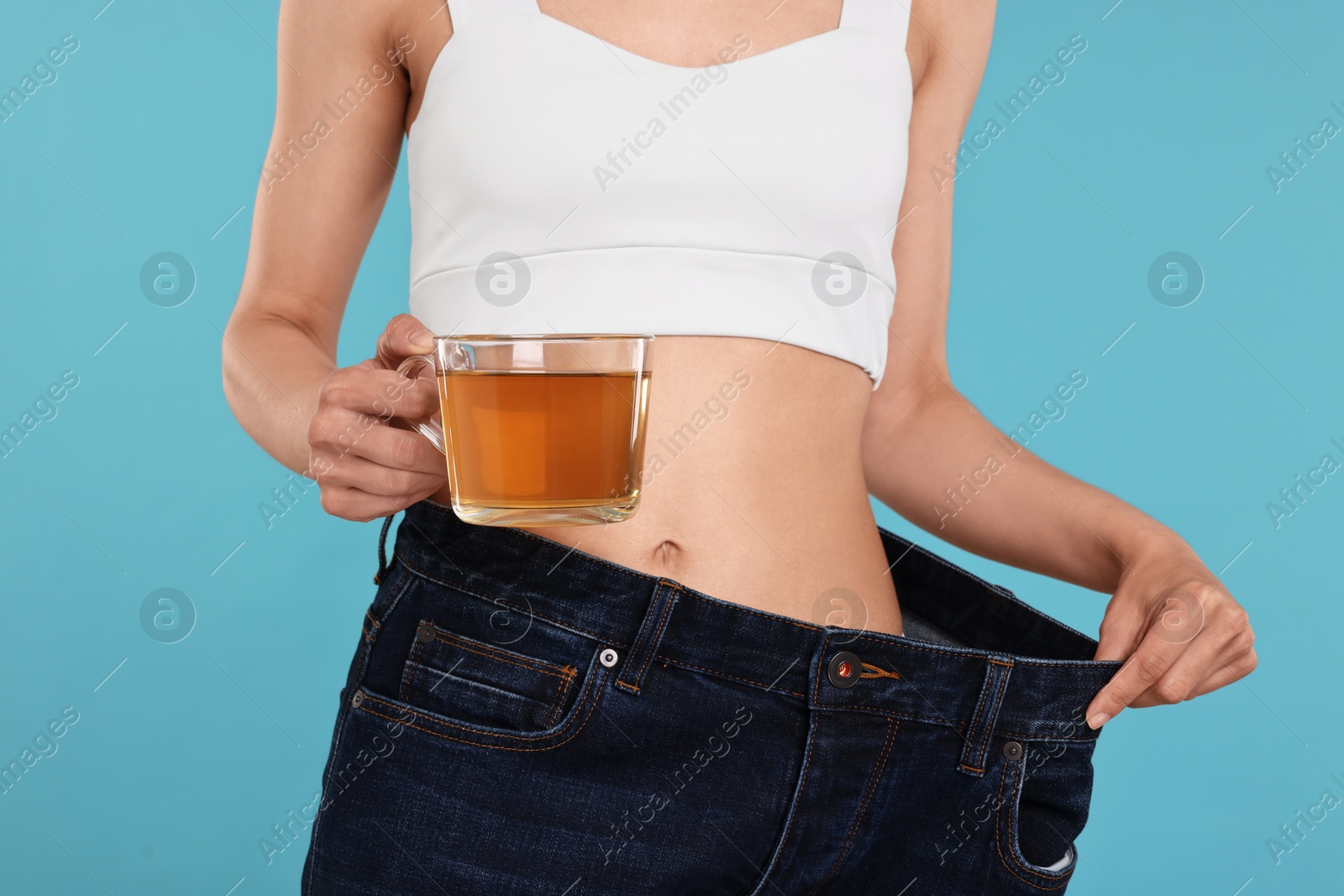 Photo of Weight loss concept. Woman with cup of diet tea and big pants on light blue background, closeup