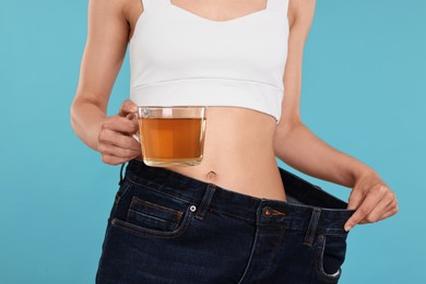 Photo of Weight loss concept. Woman with cup of diet tea and big pants on light blue background, closeup