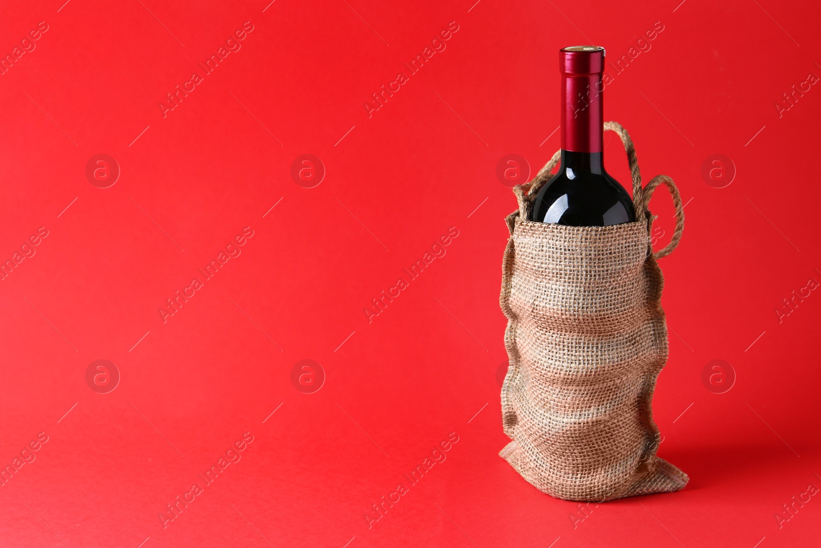 Photo of Wine bottle in burlap bag on red background, space for text