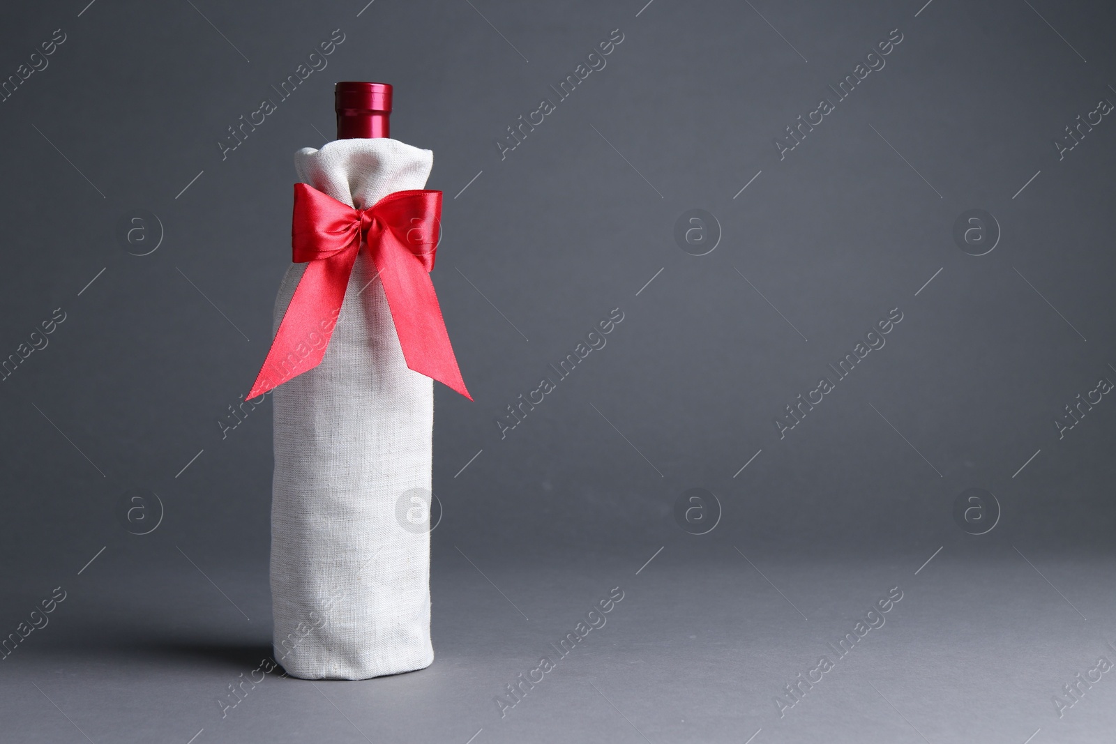 Photo of Wine bottle in burlap package with red ribbon on grey background, space for text