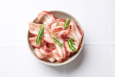 Photo of Slices of raw bacon and rosemary in bowl on white tiled table, top view