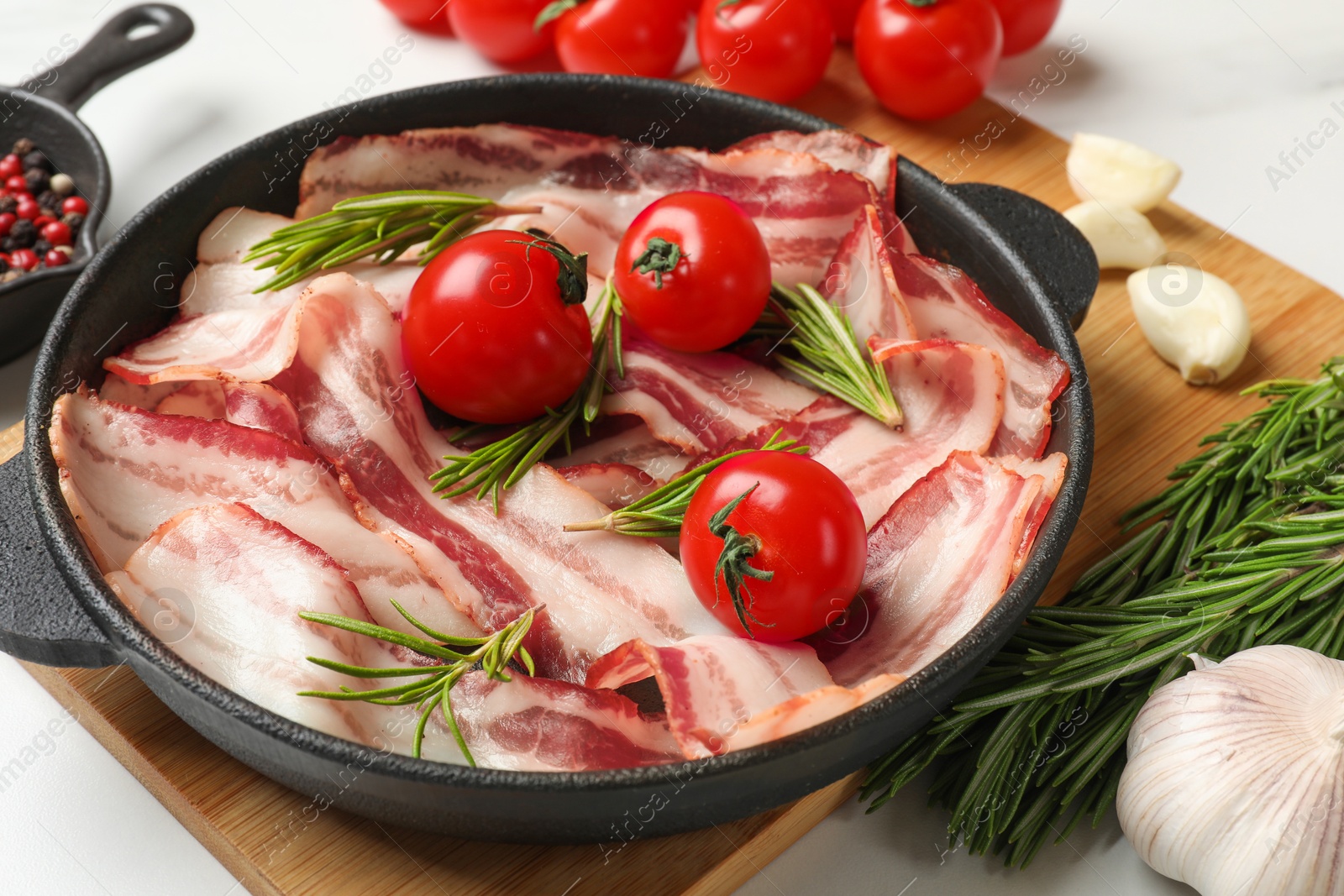 Photo of Delicious bacon slices in frying pan, spices and tomatoes on white table, closeup