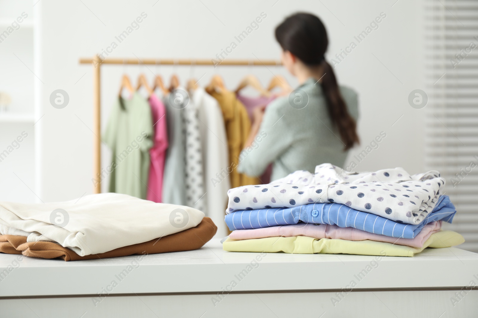 Photo of Woman choosing outfit near rack indoors, focus on stacks of clothes