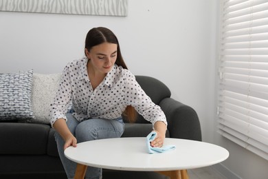 Photo of Beautiful young woman wiping coffee table with rag at home