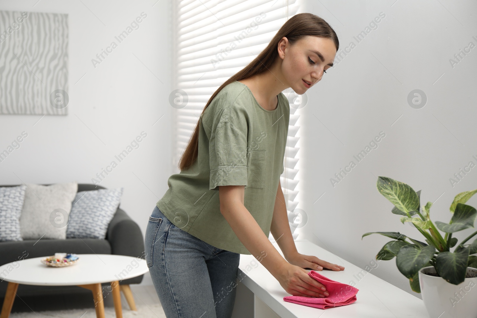 Photo of Beautiful young woman wiping furniture with rag at home