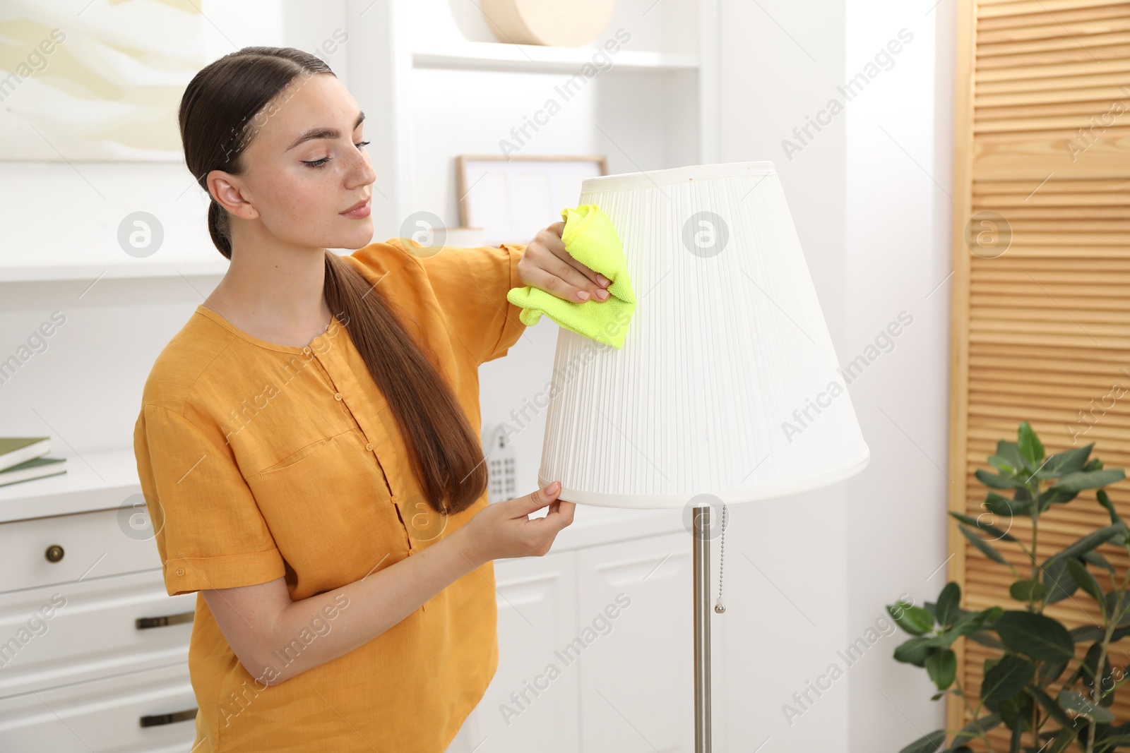 Photo of Beautiful young woman cleaning floor lamp with rag at home