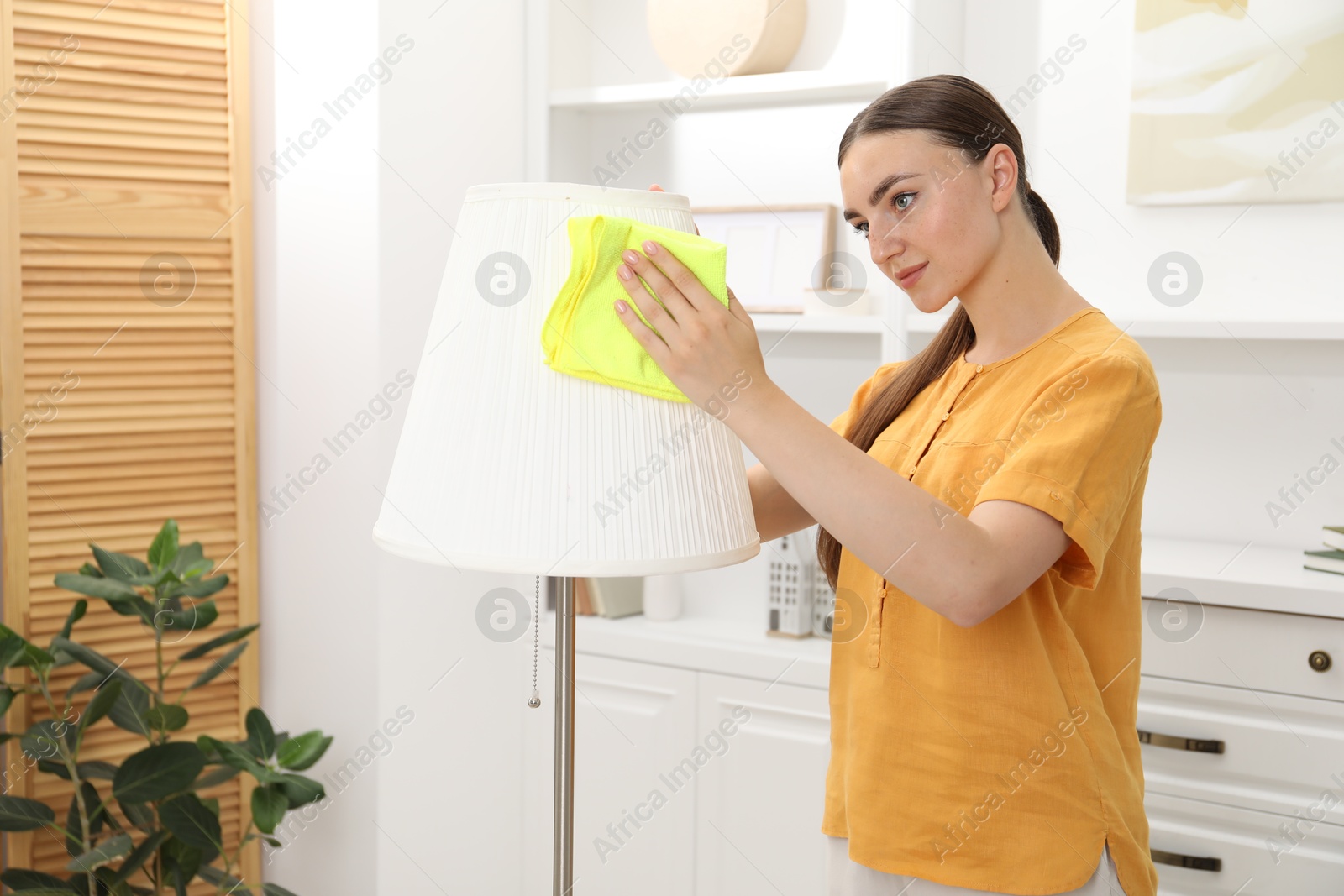 Photo of Beautiful young woman cleaning floor lamp with rag at home