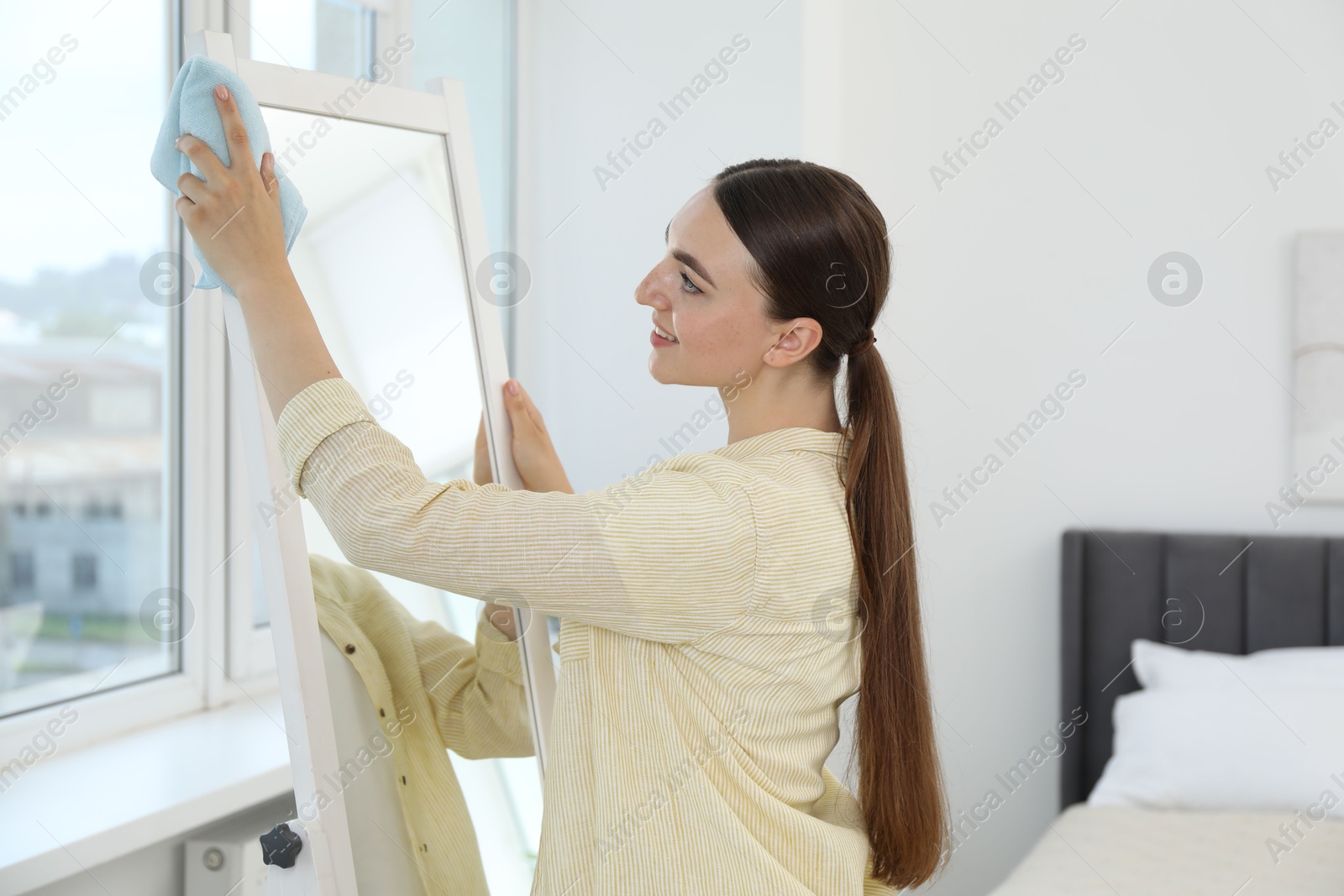 Photo of Beautiful young woman cleaning mirror with rag at home