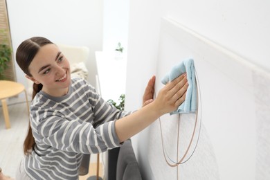 Photo of Beautiful young woman wiping picture with rag at home