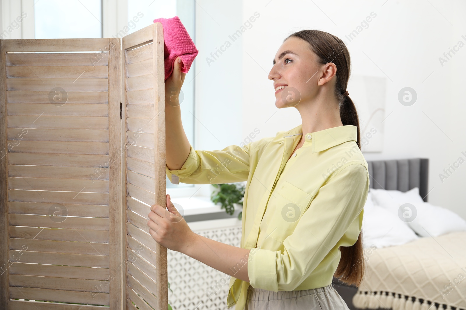 Photo of Beautiful young woman wiping folding screen with rag in bedroom