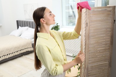 Photo of Beautiful young woman wiping folding screen with rag in bedroom