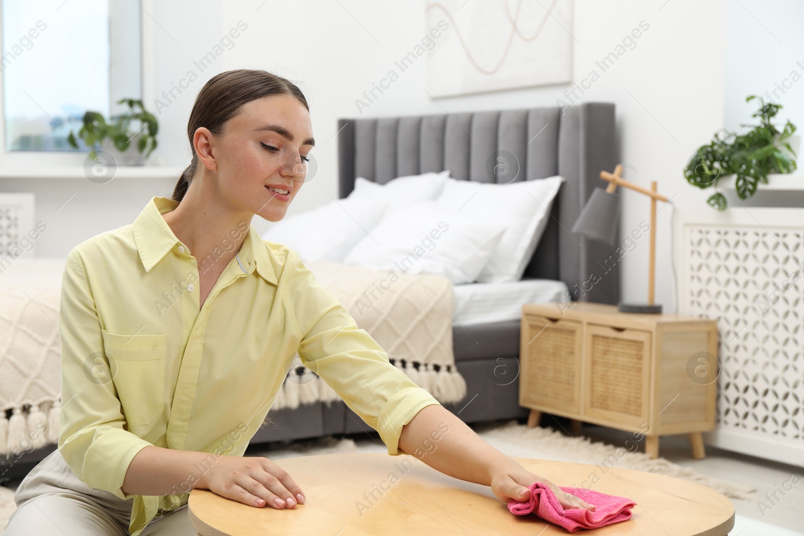 Photo of Beautiful young woman wiping coffee table with rag in bedroom