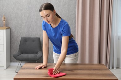 Photo of Beautiful young woman wiping wooden table with rag at home