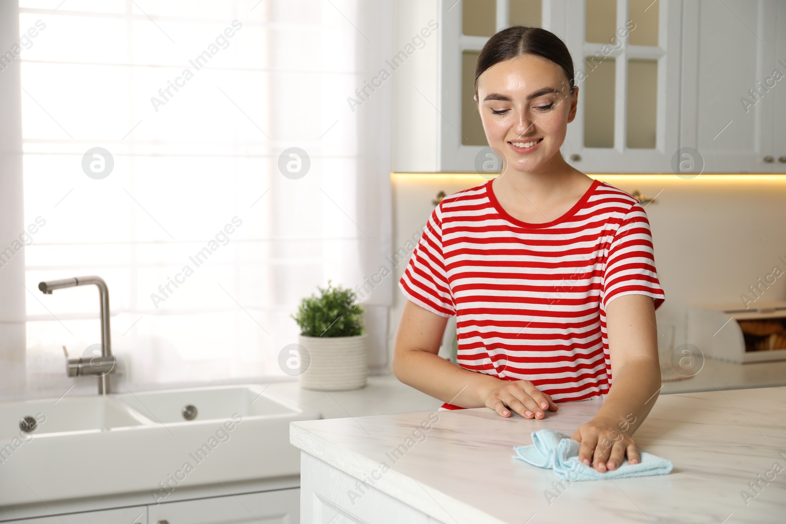 Photo of Beautiful young woman cleaning white marble table with rag in kitchen