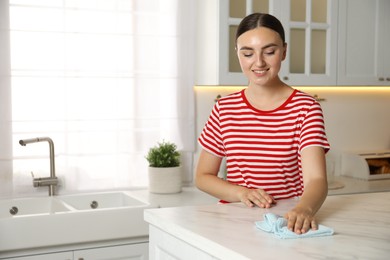 Photo of Beautiful young woman cleaning white marble table with rag in kitchen