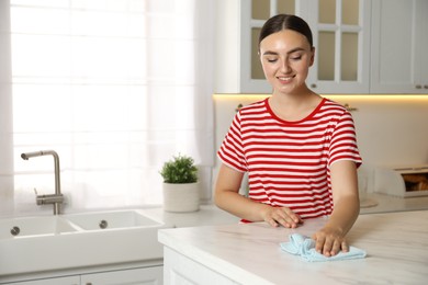 Beautiful young woman cleaning white marble table with rag in kitchen