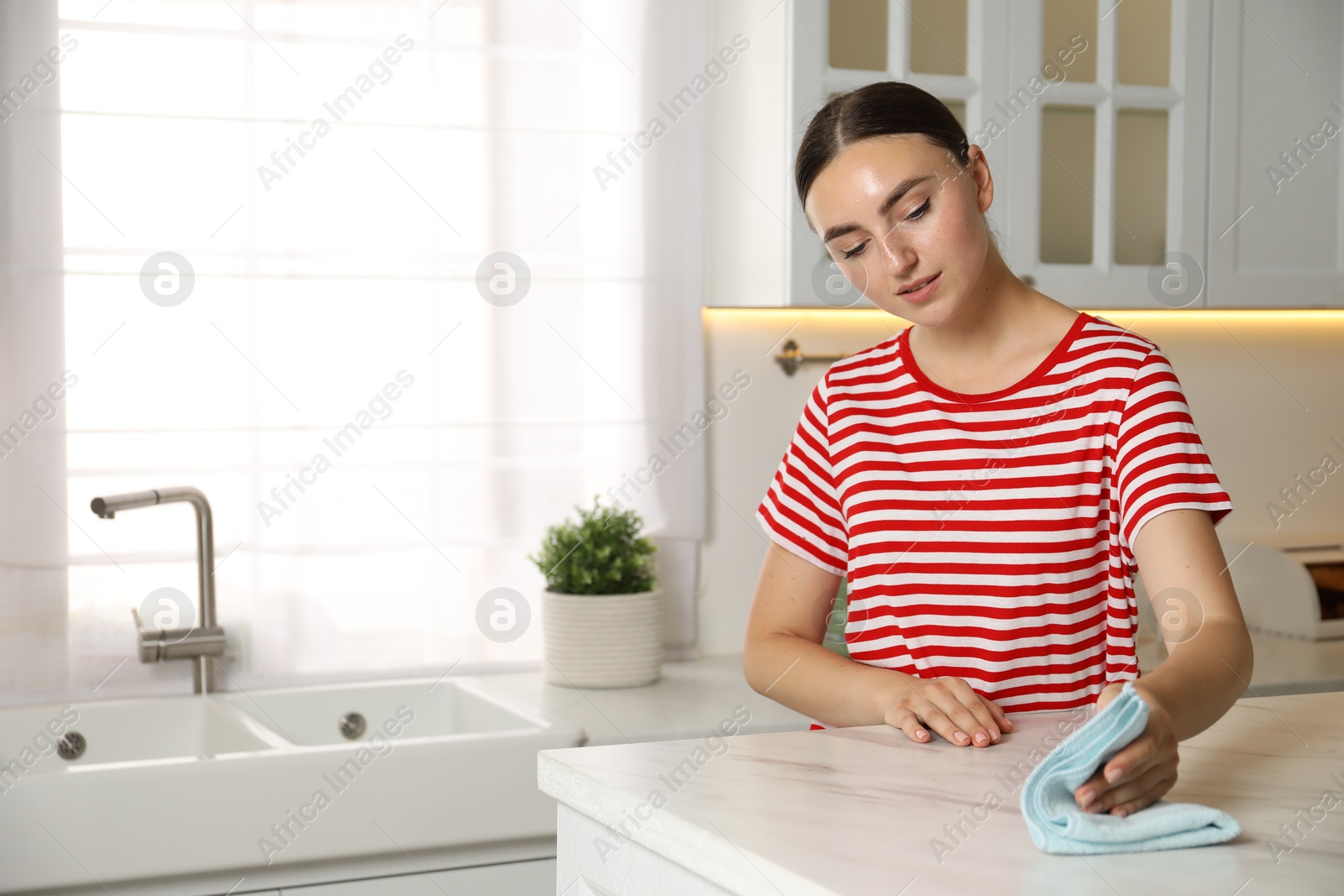 Photo of Beautiful young woman cleaning white marble table with rag in kitchen