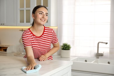 Beautiful young woman cleaning white marble table with rag in kitchen