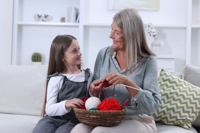 Smiling grandmother teaching her granddaughter to knit on sofa at home