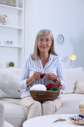 Photo of Senior woman with basket of yarn knitting on sofa at home