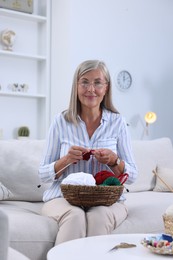 Senior woman with basket of yarn knitting on sofa at home