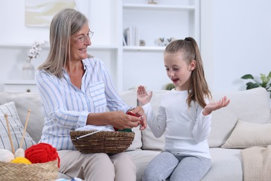 Smiling grandmother teaching her granddaughter to knit on sofa at home