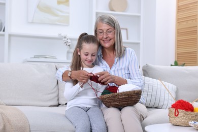 Smiling grandmother teaching her granddaughter to knit on sofa at home