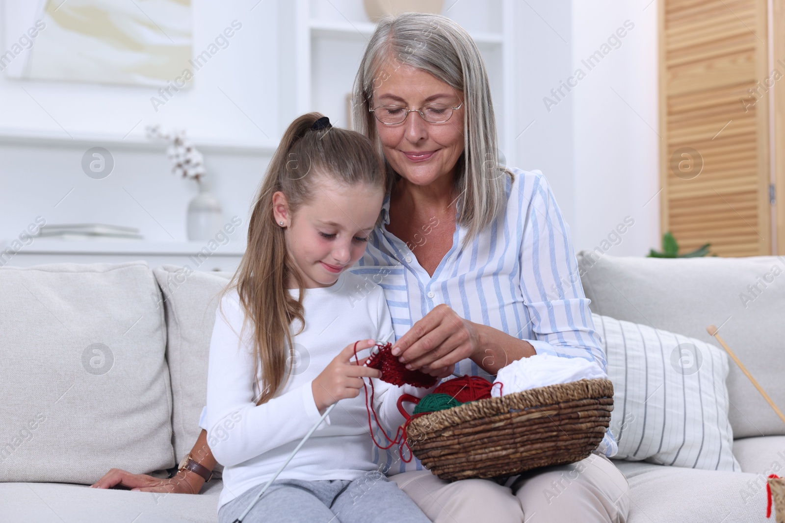 Photo of Grandmother teaching her granddaughter to knit on sofa at home