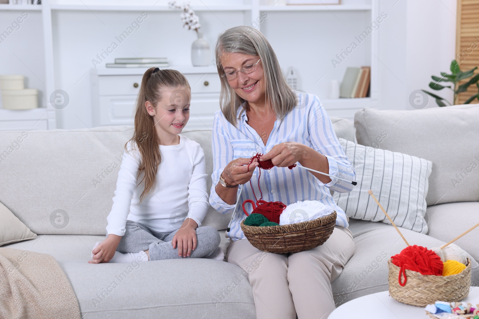 Photo of Smiling grandmother teaching her granddaughter to knit on sofa at home