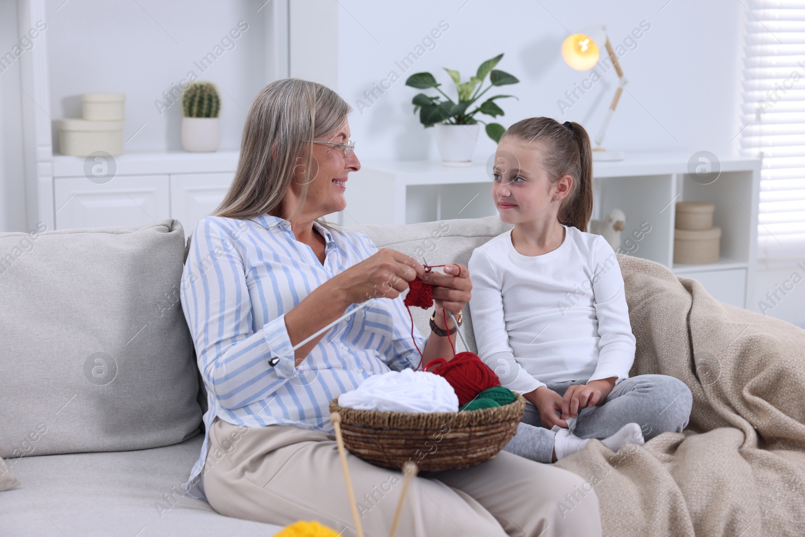 Photo of Smiling grandmother teaching her granddaughter to knit on sofa at home