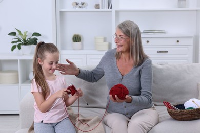 Smiling grandmother teaching her granddaughter to knit on sofa at home