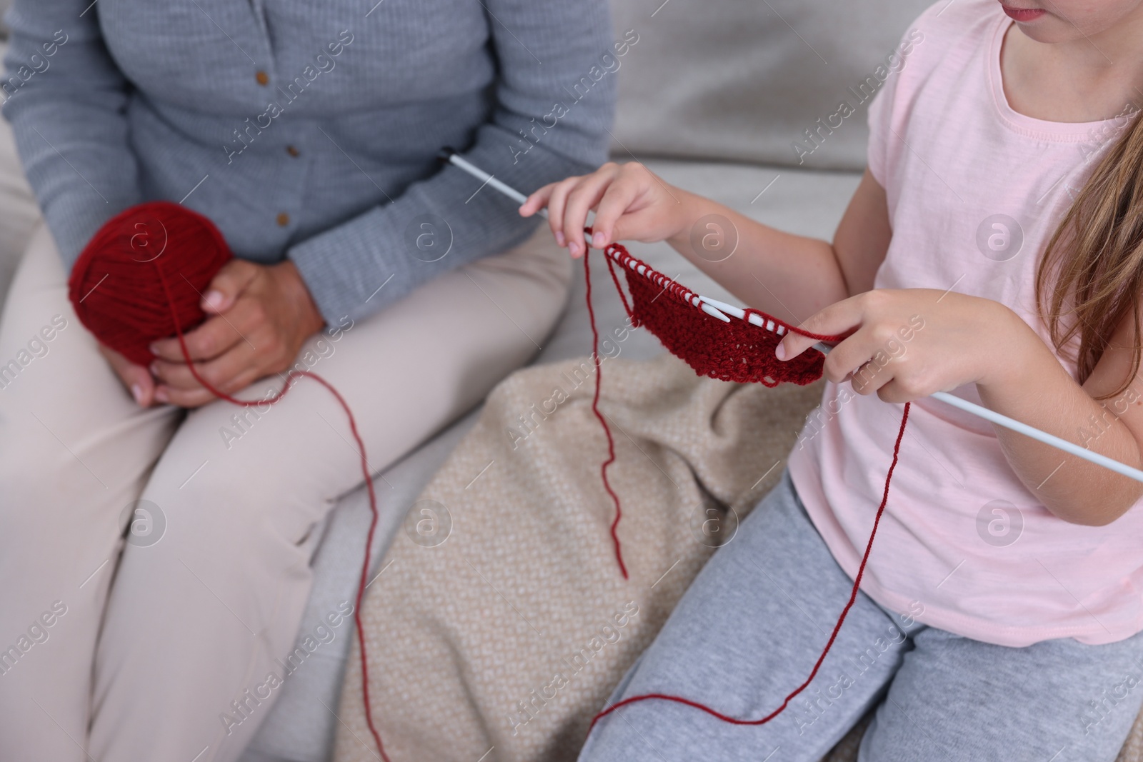 Photo of Little girl learning to knit while her grandmother holding yarn on sofa, closeup