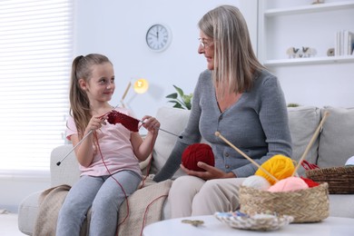 Smiling grandmother teaching her granddaughter to knit on sofa at home