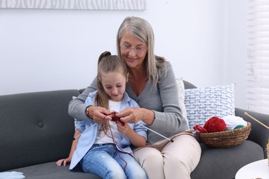 Smiling grandmother teaching her granddaughter to knit on sofa at home