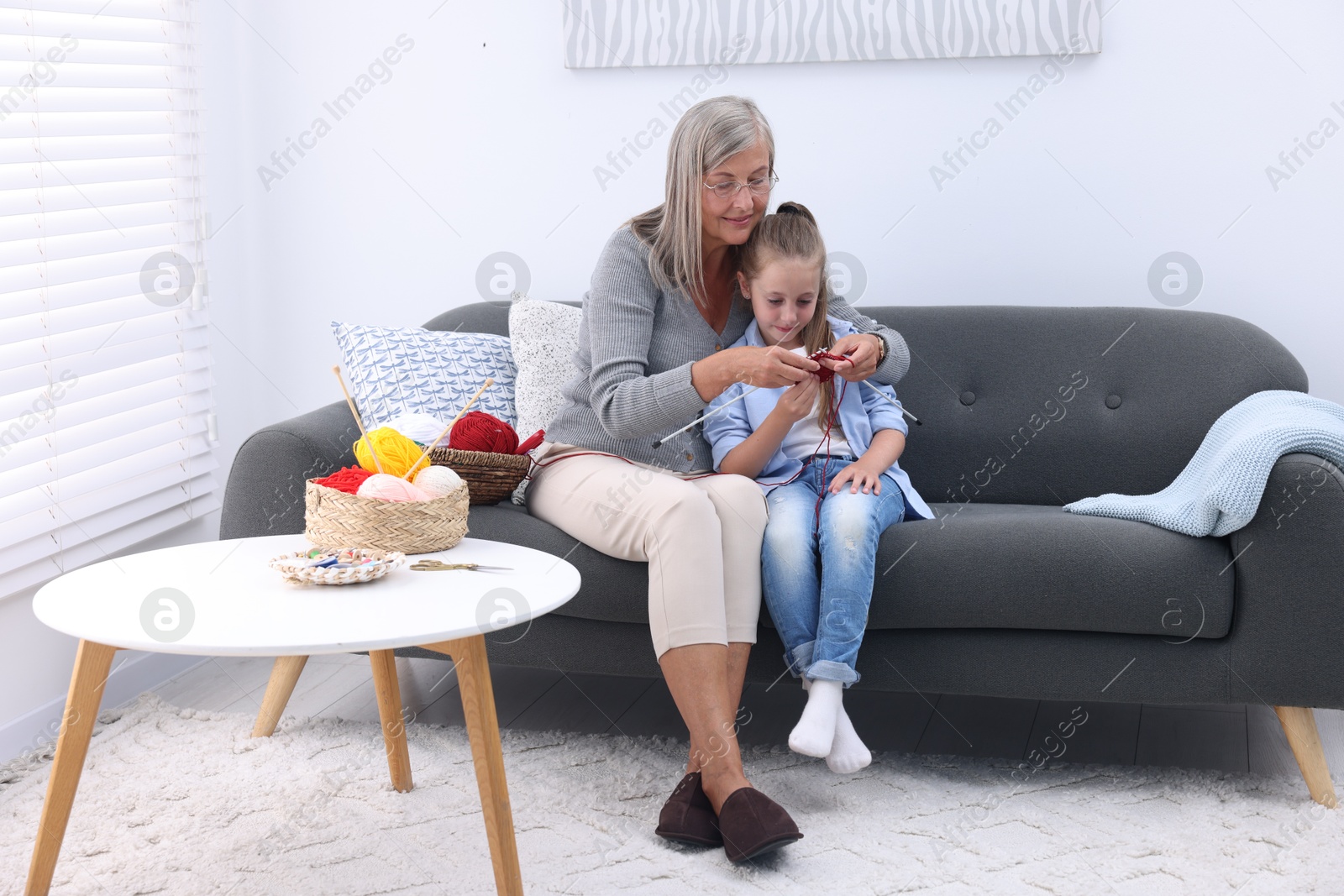 Photo of Grandmother teaching her granddaughter to knit on sofa at home