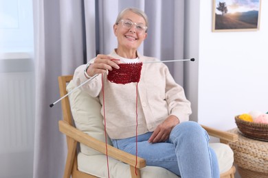 Smiling senior woman showing knitting needles on armchair at home