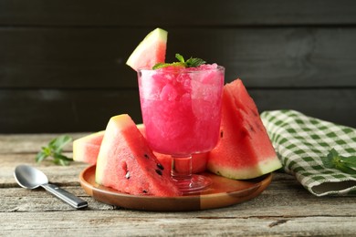 Photo of Tasty watermelon sorbet in glass dessert bowl, fresh fruit, mint and spoon on wooden table