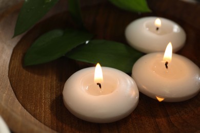 Photo of Burning candles and green leaves in water on table, closeup