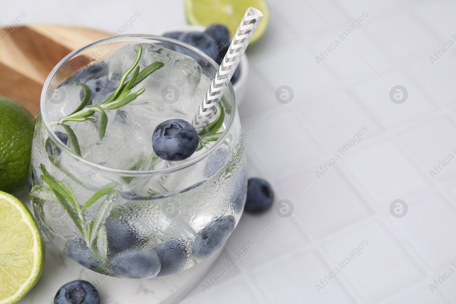 Photo of Refreshing water with blueberries and rosemary in glass on white tiled table, closeup. Space for text