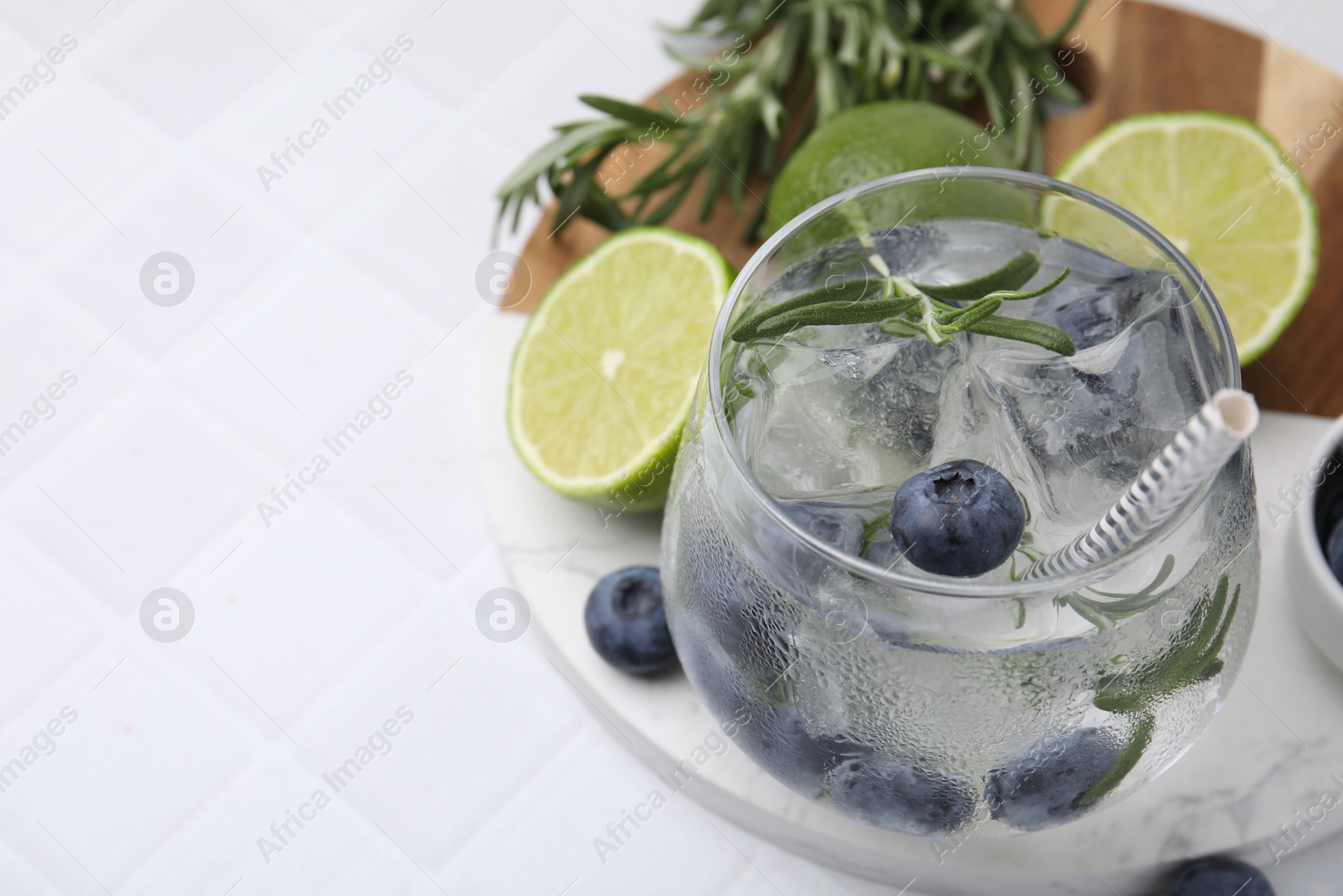 Photo of Refreshing water with blueberries and rosemary in glass on white tiled table, closeup. Space for text