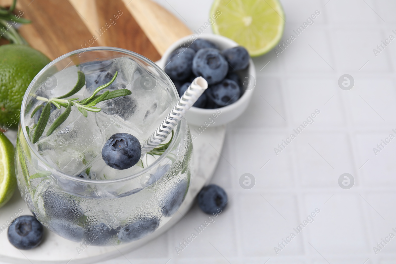 Photo of Refreshing water with blueberries and rosemary in glass on white tiled table, closeup. Space for text