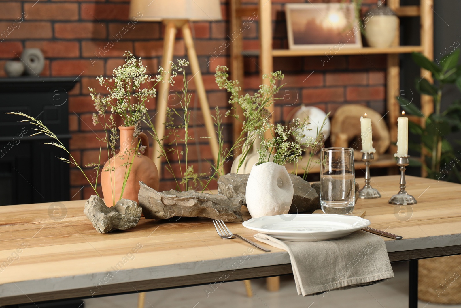 Photo of Set of clean dishware, vases with beautiful flowers, stones and burning candles on wooden table in stylish dining room