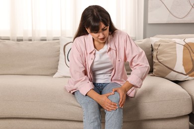 Upset woman suffering from knee pain on sofa at home