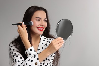 Smiling woman with mirror applying makeup on light grey background