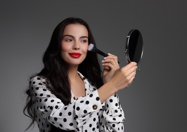 Beautiful woman with mirror applying makeup on grey background