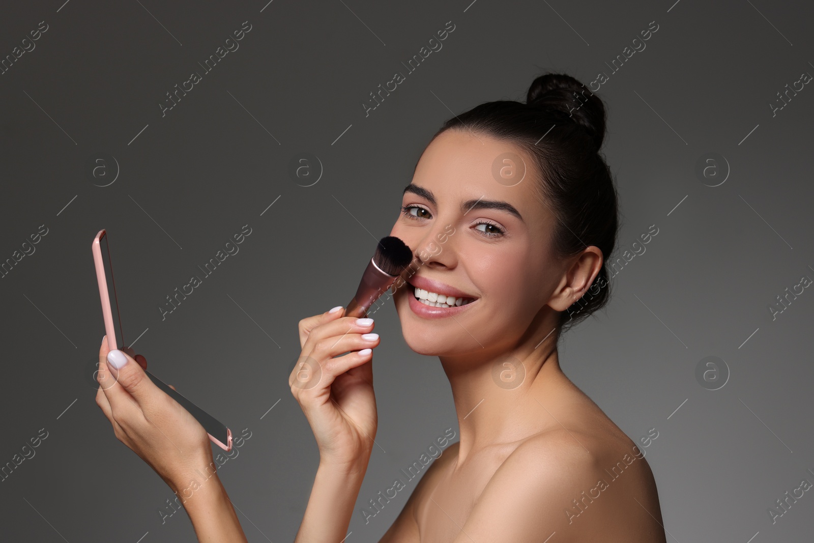 Photo of Smiling woman with mirror applying makeup on grey background